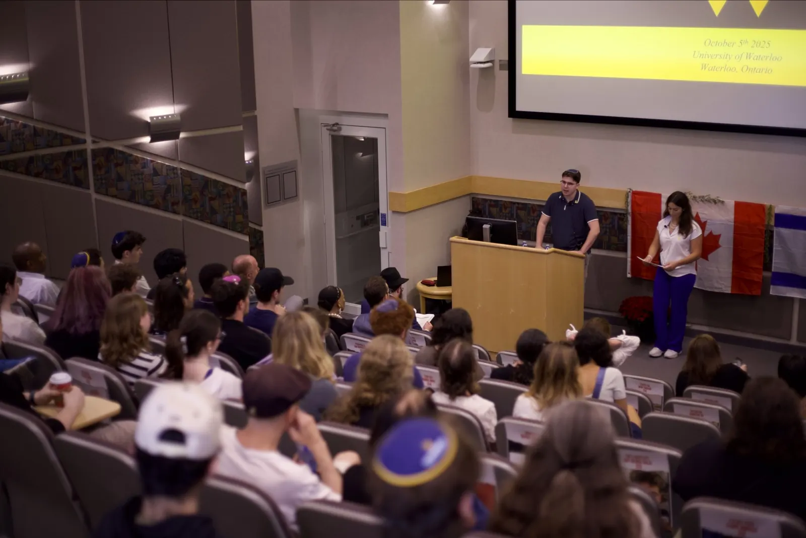 A packed lecture hall at the University of Waterloo’s October 7th commemoration, flags flanking the speakers